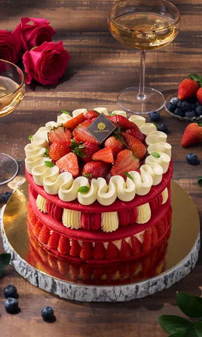 Macaron Strawberry Cake topped with fresh strawberries and cream, styled with roses and champagne glasses on a wooden table.