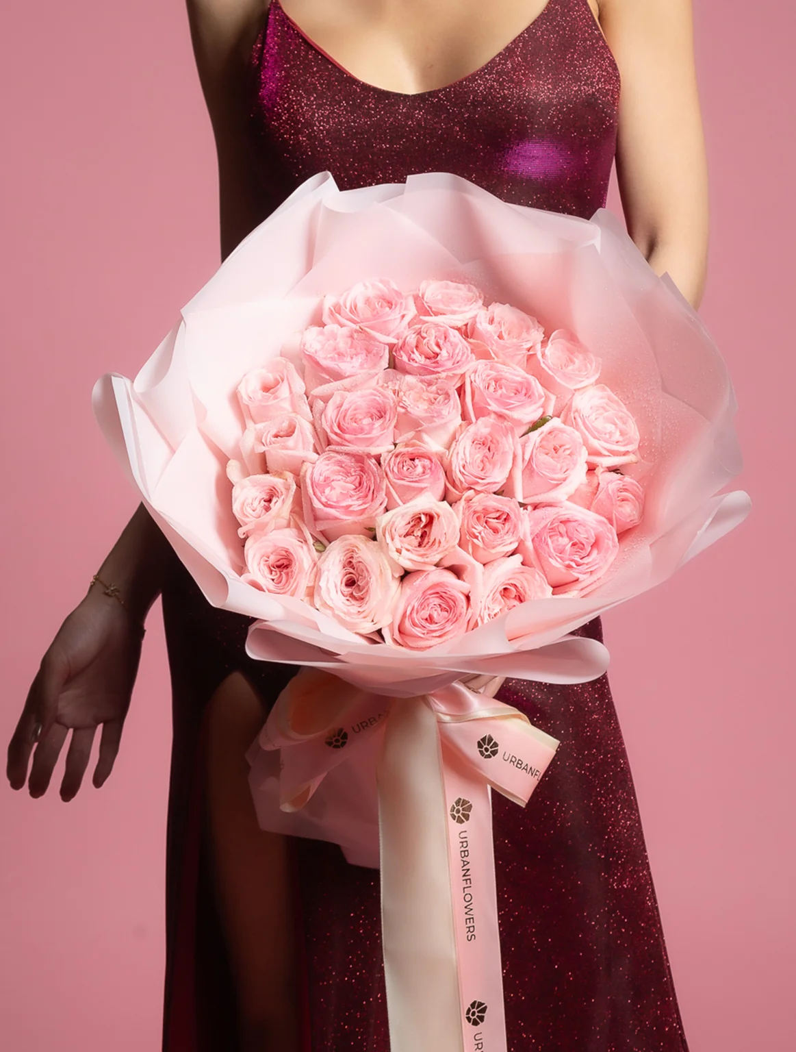 A woman in a deep red dress holds an extravagant bouquet of pink roses wrapped in elegant white paper, set against a soft pink background — a romantic and luxurious floral arrangement perfect for special occasions.