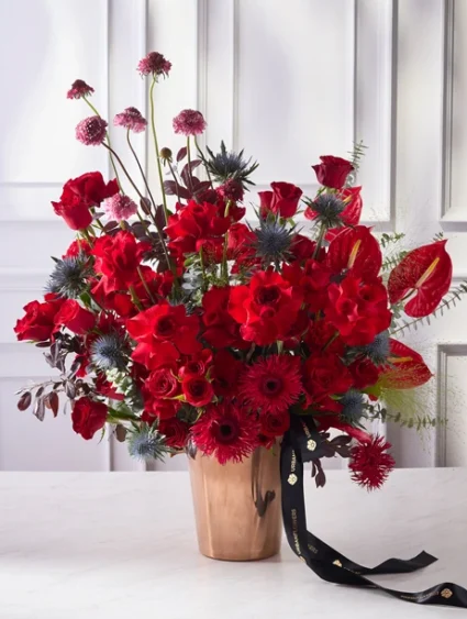 ช่อดอกไม้แชมเปญสุดหรู ดอกกุหลาบและเยอบีร่าสีแดงจัดคู่กับหน้าวัวในแจกันสีบรอนซ์ เหมาะสำหรับงานฉลองสุดหรู